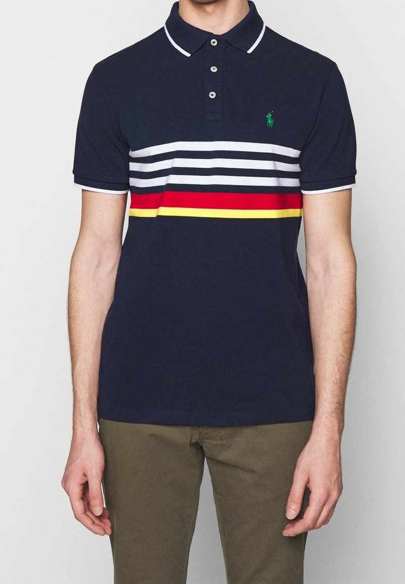 Homme portant un polo bleu marine avec des rayures horizontales blanches, rouges et jaunes et un pantalon vert olive, debout, les bras détendus.