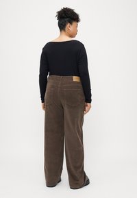 Brown corduroy wide-leg pants with a textured finish, featuring rear pockets and a small tan label on the waistband. Black long-sleeve top.