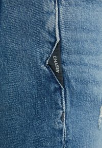 Šviesiai mėlyni džinsai su tekstūruotu paviršiumi, nežymiu blukimu ir juodu etikete su balta užrašu, siūtu į siūlę.