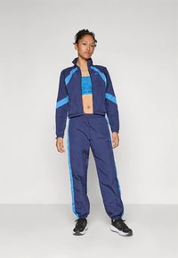 Blå og marineblå joggedress med en glidelåsjakke med blå striper, matchende bukser og en teksturert blå topp. Svarte treningssko.