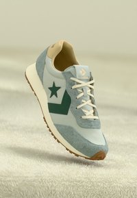 Light blue sneaker featuring gray suede accents, a green star logo, and beige laces; rubber sole with brown tread pattern.