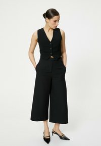 Zwarte mouwloze vest met een V-halslijn en drie knopen, gecombineerd met zwarte wijde broek en glanzende zwarte slingbackhakken.