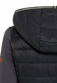 Veste noire matelassée à capuche avec des surpiqûres rembourrées dans le dos et des manches en tissu gris foncé avec un petit patch marron sur le bras gauche.