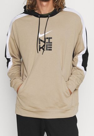 Beige Nike hættetrøje med sort/hvide striber på ærmerne, sorte snorer og vertikalt Nike-logo på brystet, båret med hænderne i lommerne.