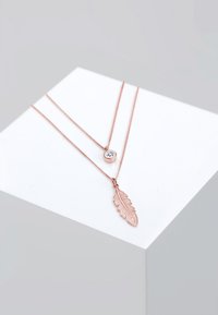 Roségoldene, geschichtete Halsketten mit einem zarten Federanhänger und einem runden Kristallakzent, präsentiert auf einer hellgrauen geometrischen Oberfläche.