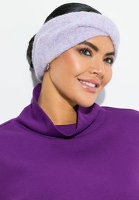 Fascia per la testa lavorata a maglia in tonalità pastello di viola e rosa, caratterizzata da un design attorcigliato. Il modello indossa un top viola a collo alto e a coste.