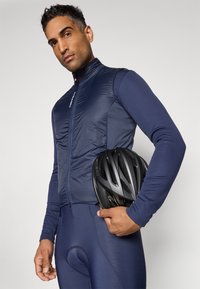 Chaleco de ciclismo azul marino con cuello alto, diseño ajustado y cierre de cremallera; sosteniendo un casco negro. Textura suave, minimalista en marcas.