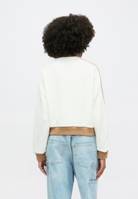 Sudadera blanca corta con puños y dobladillo marrones acanalados, que presenta un corte relajado y una textura suave, mostrada desde la parte posterior.