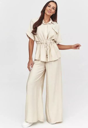 Kobieta z długimi brązowymi włosami, ubrana w beżowe spodnie o szerokich nogawkach oraz pasującą krótką koszulę na guziki ze ściągaczem w talii, stoi swobodnie.