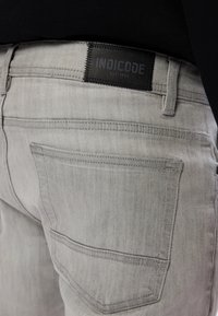 Svijetlosivi traperice s crnim kožnim detaljem s oznakom "INDICODE EST 1993". Prikazuje stražnji džep i detalje šivanja.