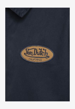 Écusson du logo Von Dutch California en fil orange et noir sur tissu bleu foncé.