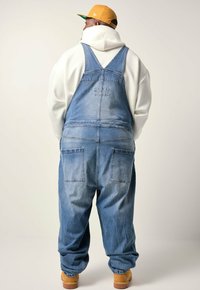 Mand med ryggen til iført en hvid hoodie, løse denim overalls, gule støvler og en sennepsgul baseball cap foran en ensfarvet baggrund.