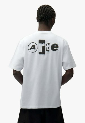 Persona con capelli intrecciati che indossa una maglietta bianca con lettere nere in font misto che formano la parola "Arte" sulla schiena, in piedi davanti a uno sfondo semplice.