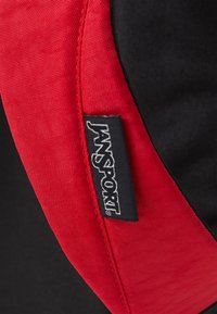 Coin de sac à dos en tissu noir et rouge avec un matériau texturé ; présente une étiquette de logo Jansport visible cousue sur le bord.