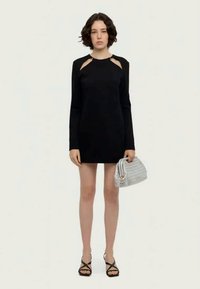 Vestido mini preto de manga longa com recortes nos ombros, textura suave, acompanhado de uma bolsa prateada franzida e sandálias pretas com tiras.