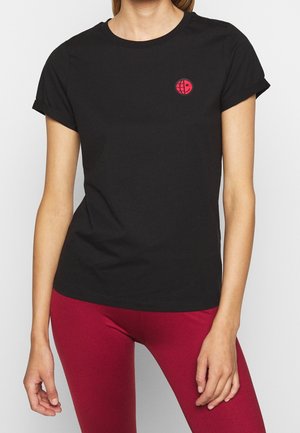 Femme portant un t-shirt noir uni à manches courtes avec un petit logo circulaire rouge et noir sur la poitrine gauche, associé à un pantalon rouge.