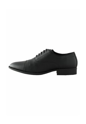 Zapato de vestir de cuero negro con textura suave, punta redondeada y tacón bajo, que cuenta con seis ojetes para los cordones y sutiles detalles de costura.