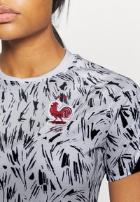 Camisa atlética gris con un patrón texturizado negro, cuello redondo y un emblema de gallo rojo con estrellas y "FFF" bordado debajo.