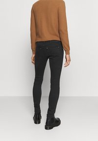 Person i smala svarta jeans, svarta stövlar och en brun långärmad topp som står vänd mot en enkel ljusgrå bakgrund.