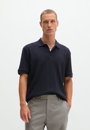 Man draagt een donkerblauw poloshirt met korte mouwen en lichtgrijze broek, staand tegen een effen witte achtergrond.