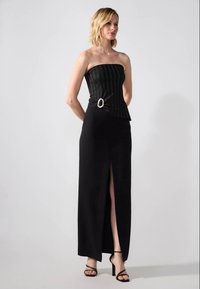 Woman wearing a strapless black dress with a side slit and buckle detail, paired with black strappy high-heeled sandals, standing against a gray background.