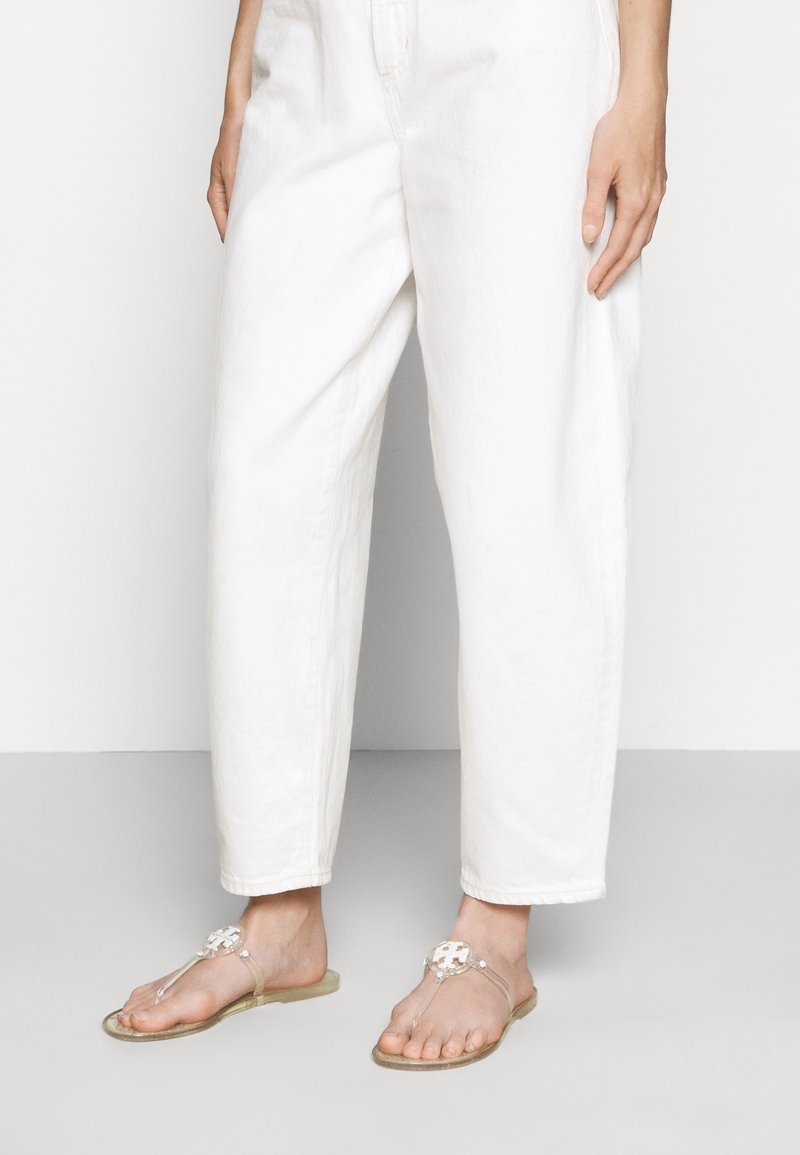 Personne portant un pantalon ample blanc taille haute et des sandales plates transparentes avec un emblème décoratif blanc sur les brides.