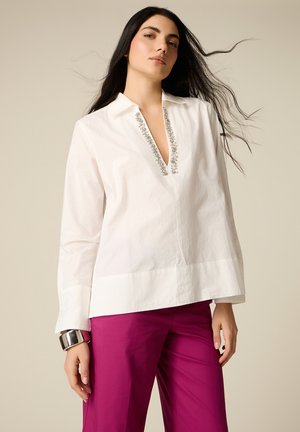 Pitkätukkainen nainen, jolla on valkoinen helmillä koristeltu V-kauluksinen paita ja magentanväriset housut, seisoo yksivärisen beige taustan edessä.