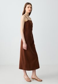 Abito midi in lino marrone con spalline sottili, corpino leggermente arricciato e orlo dritto. Abbinato a sandali con tacco nude.