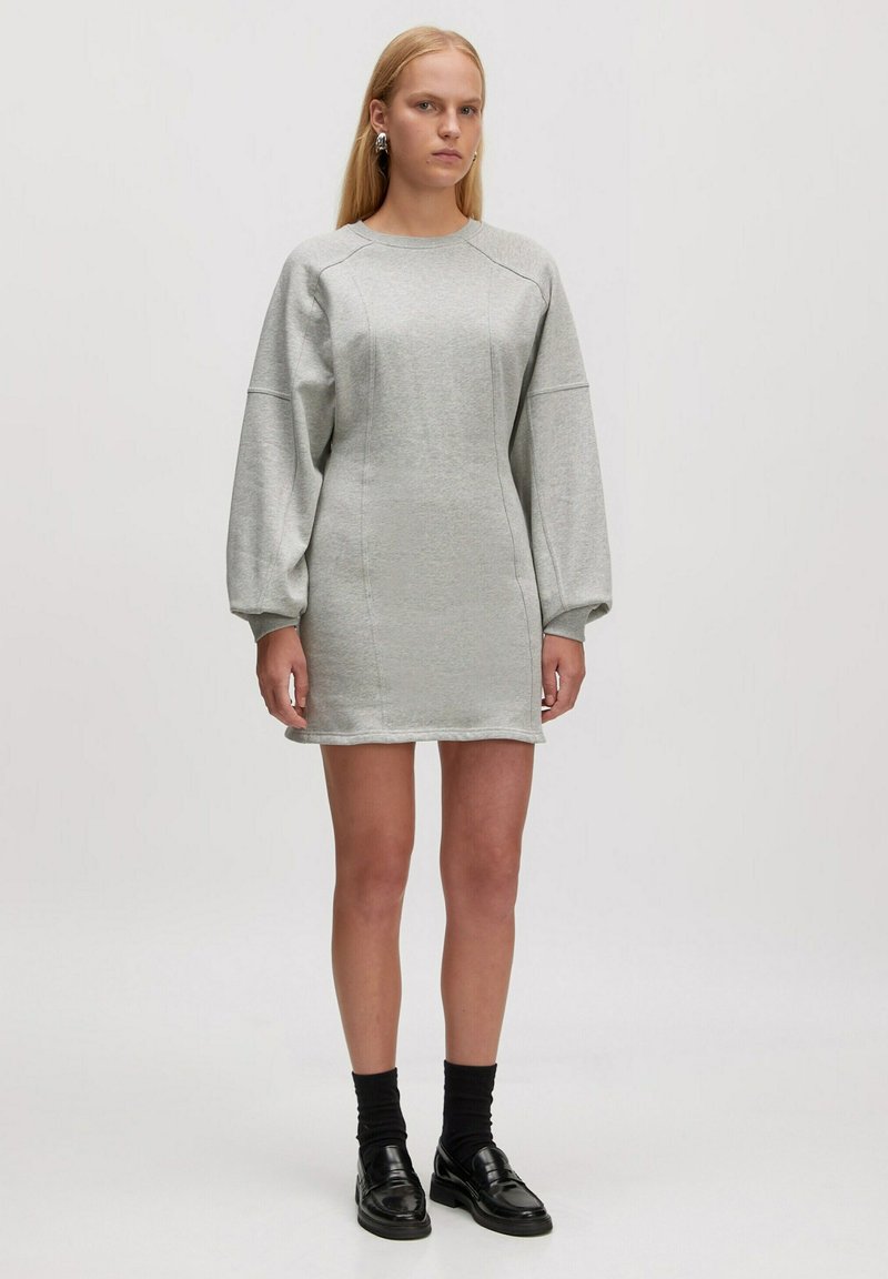 Robe en sweat gris à manches longues, texture douce, silhouette ajustée et détails cousus ; assortie à des chaussettes noires et des mocassins.