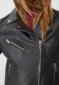 Černá kožená motocyklová bunda se stříbrnými zipy, patentkami a několika zipovými kapsami, s texturovaným povrchem a přizpůsobeným střihem.