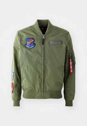 Olivgrüne Bomberjacke mit gerippten Bündchen, Reißverschluss vorne und bestickten Aufnähern, darunter ein Abzeichen und rotes Akzentband am Ärmel.