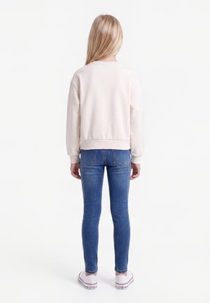 Enfant debout de dos, portant un sweat-shirt clair, un jean slim bleu et des baskets blanches sur fond blanc.