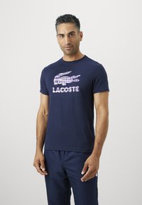 Άνδρας που φοράει ένα ναυτικό μπλε T-shirt Lacoste με μεγάλο ανοιχτό μοβ λογότυπο κροκόδειλου και ναυτικά μπλε παντελόνια μπροστά από απλό ανοιχτό φόντο.