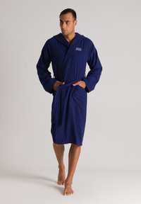 Accappatoio blu navy in terry, dotato di cappuccio, tasche frontali e cintura da annodare. Design semplice con una superficie testurizzata.