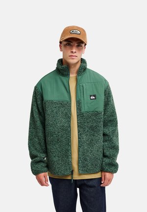 Jeune homme portant une veste polaire verte avec une poche poitrine matelassée et une casquette de baseball marron, debout sur un fond uni.