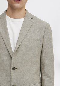 Ljus beige linnety blazer med en subtil textur, med klassiska slag, en knappstängning och en bröstficka. Bärs över en vit T-shirt med rund hals.