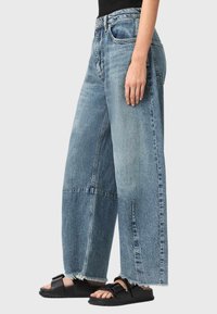Vaqueros de mezclilla azul claro de pierna ancha con dobladillos desflecados. Presentan una cintura alta, bolsillos clásicos y un ajuste holgado. Combinados con sandalias negras.