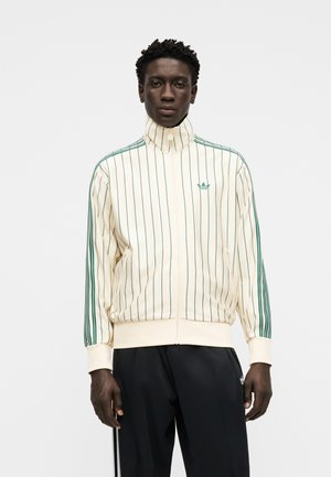 Hombre con chaqueta crema de cuello alto con rayas verticales verdes y pantalones negros a rayas verdes a juego, de pie contra un fondo liso.