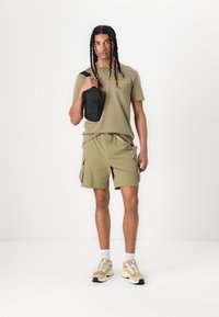 Olijfgroene T-shirt met de tekst "OUTSIDER", bijpassende shorts en beige sneakers. Zwarte tas als schoudertas, minimalistisch ontwerp.