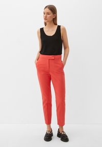 Zwarte mouwloze top en koraalkleurige tapered broeken met vo pockets, gladde textuur; gecombineerd met zwarte loafers met een decoratieve ketting.