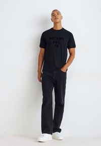 Svart kortermet t-skjorte med "NOTTING HILL 73" trykt i mørke bokstaver, kombinert med svarte jeans og hvite joggesko.