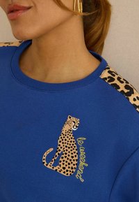 Blauwe sweatshirt met een luipaardprint op de voorkant en luipaardprint accenten op de schouders. Zachte textuur, ronde hals en gouden letters.
