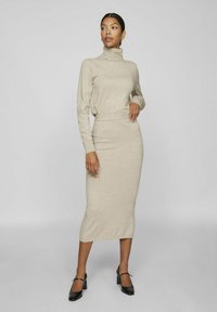 Suéter de punto beige con cuello alto, combinado con una falda midi ajustada a juego. La modelo lleva unos zapatos de tacón negros, que exhiben un diseño simple y elegante.