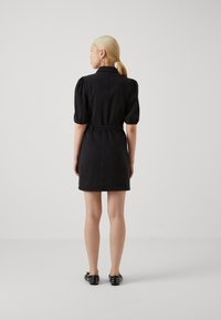 Robe en denim noir avec des manches bouffantes et une taille ceinturée, présentant une coupe droite et un ourlet plat, portée avec des chaussures noires à clous.