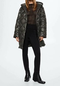 Donna con cappotto imbottito grigio scuro con cappuccio, top trasparente a righe nere, pantaloni neri aderenti e stivali neri con tacco, su sfondo neutro.