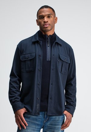 Navy blue button-up shirt with two chest pockets, made of soft material, layered over a dark knitted pullover with a zip neck.