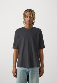 Mørk grå bomullst-skjorte med rund hals og kort ärm, vist sammen med løse lyseblå jeans.