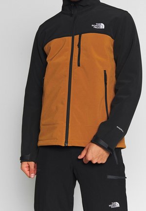 Homme portant une veste zippée marron et noire et un pantalon noir avec les logos The North Face et des poches zippées.