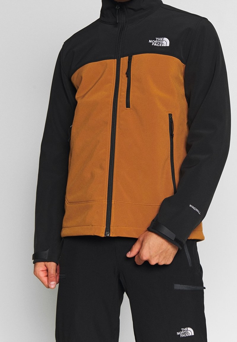 Homme portant une veste zippée marron et noire et un pantalon noir avec les logos The North Face et des poches zippées.