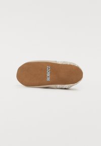 Beige fottøy med en myk semsket såle og stoffoverdel med blomstermønster. "Robeez"-logoen i blått plassert nær hælen på en glatt overflate.